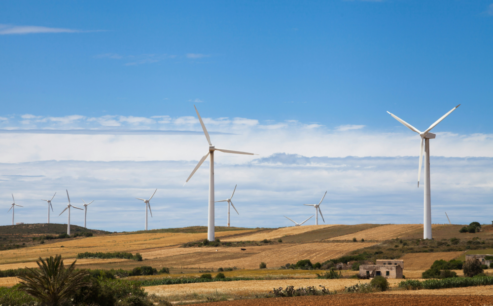 Wind turbines in a rural landscape with fields, hills, and scattered buildings under a partly cloudy blue sky.