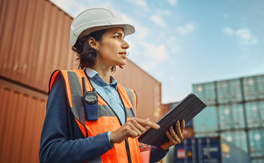 Female engineer or logistics worker in safety gear using a tablet at a shipping container yard.